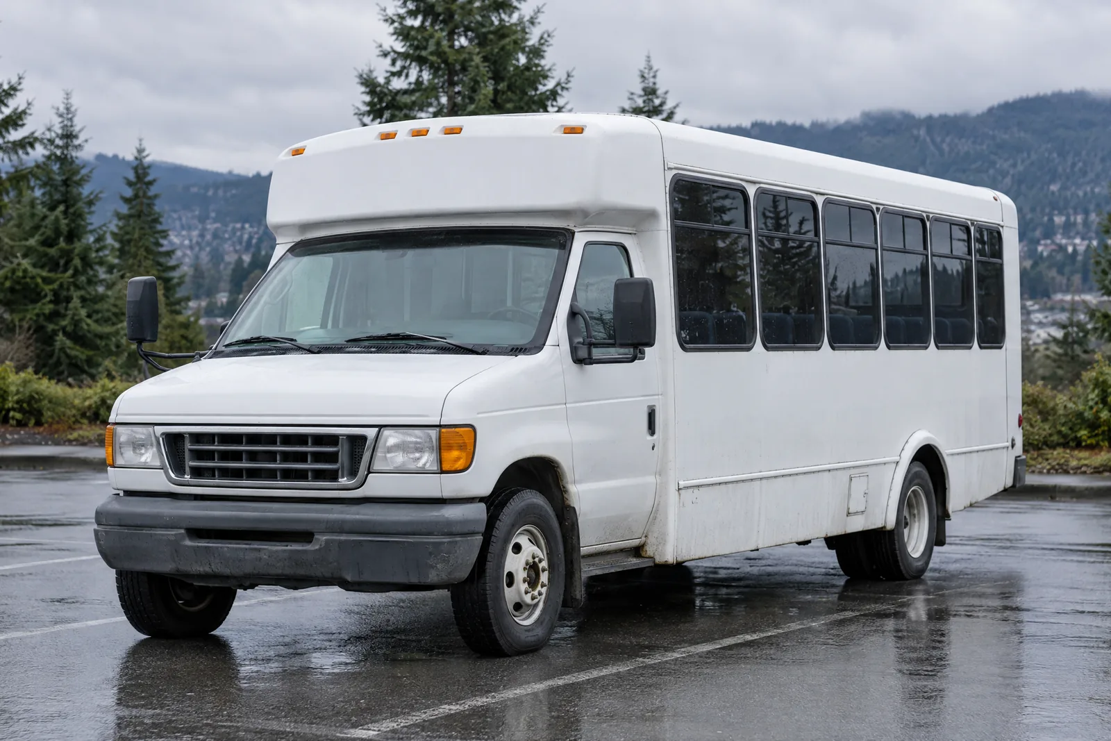 Plain white shuttle bus parked on a wet parking lot with evergreen trees and hills behind